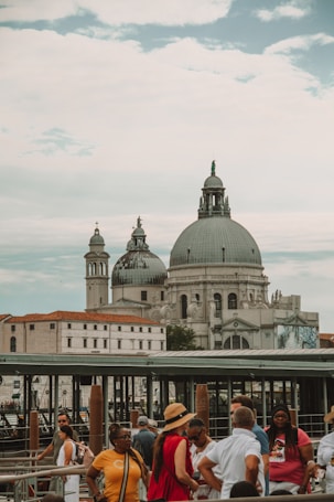 A group of people gathered near a waterfront with a historic architectural structure in the background, featuring large domes and intricate details. The sky is partly cloudy, adding to the grandeur of the scene. The area appears to be a popular tourist spot with a bustling atmosphere.