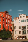 Two adjacent buildings with distinct Mediterranean architecture, one painted in vibrant red and the other in pastel cream. Green shutters adorn the windows, and a large green tree stands in front of the red building. A group of people are seated at a small outdoor cafe under an umbrella.