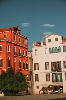 Two adjacent buildings with distinct Mediterranean architecture, one painted in vibrant red and the other in pastel cream. Green shutters adorn the windows, and a large green tree stands in front of the red building. A group of people are seated at a small outdoor cafe under an umbrella.