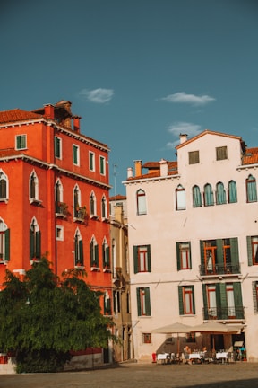 Two adjacent buildings with distinct Mediterranean architecture, one painted in vibrant red and the other in pastel cream. Green shutters adorn the windows, and a large green tree stands in front of the red building. A group of people are seated at a small outdoor cafe under an umbrella.