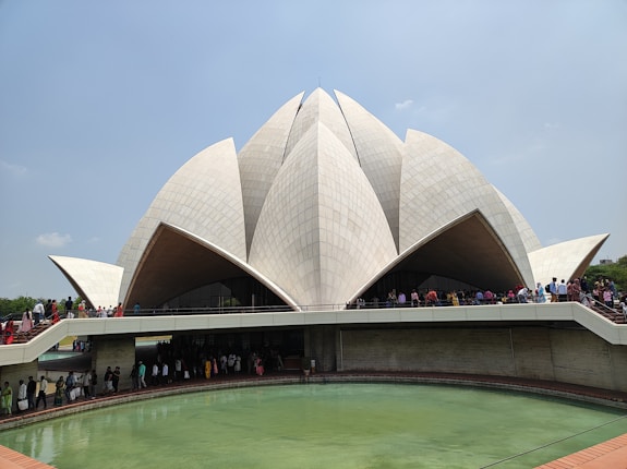 A large, architecturally unique building resembling a lotus flower with white petal-like structures surrounding a central dome. The structure is elevated above a pool of water and numerous people are seen walking around it, indicating a popular and likely significant tourist attraction.