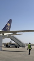 An airport scene featuring a parked airplane with its tail in view, displaying a logo. A mobile staircase is positioned for boarding. A person wearing a high-visibility safety jacket walks nearby on the tarmac.