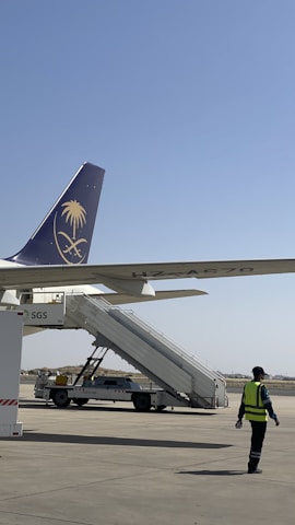 An airport scene featuring a parked airplane with its tail in view, displaying a logo. A mobile staircase is positioned for boarding. A person wearing a high-visibility safety jacket walks nearby on the tarmac.