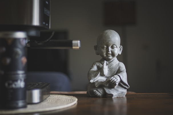 A small statue of a serene monk in a meditative pose sits on a wooden surface. The monk has a peaceful expression, eyes closed, and wears traditional robes. To the left, part of a coffee-making machine and a canister labeled 'Café' are visible, with a muted and cozy ambiance in the background.