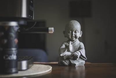 A small statue of a serene monk in a meditative pose sits on a wooden surface. The monk has a peaceful expression, eyes closed, and wears traditional robes. To the left, part of a coffee-making machine and a canister labeled 'Café' are visible, with a muted and cozy ambiance in the background.
