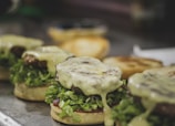 Several cheeseburgers are lined up, featuring juicy hamburger patties with melted cheese and fresh lettuce on round buns. The cheese appears to be melting, adding a glossy texture under the warm lighting.