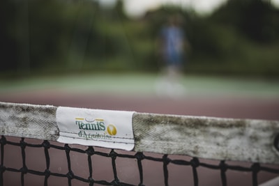 The image focuses on a close-up of a tennis net with a white band labeled 'Tennis d'Aquitaine'. The background is blurred, showing indistinct greenery and a person in sports attire, suggesting an outdoor tennis court setting.