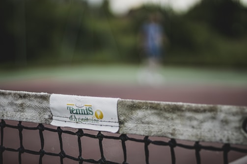 The image focuses on a close-up of a tennis net with a white band labeled 'Tennis d'Aquitaine'. The background is blurred, showing indistinct greenery and a person in sports attire, suggesting an outdoor tennis court setting.