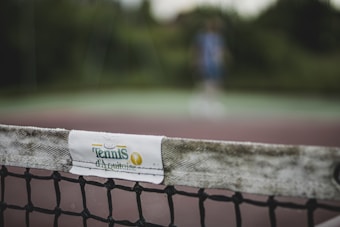 The image focuses on a close-up of a tennis net with a white band labeled 'Tennis d'Aquitaine'. The background is blurred, showing indistinct greenery and a person in sports attire, suggesting an outdoor tennis court setting.