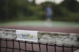 The image focuses on a close-up of a tennis net with a white band labeled 'Tennis d'Aquitaine'. The background is blurred, showing indistinct greenery and a person in sports attire, suggesting an outdoor tennis court setting.