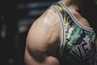 A close-up of a muscular person's shoulder wearing a floral-patterned tank top. The tank top features vivid green leaves and white flowers with subtle yellow accents. The focus is on the prominent veins and well-defined muscle on the shoulder.
