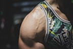 A close-up of a muscular person's shoulder wearing a floral-patterned tank top. The tank top features vivid green leaves and white flowers with subtle yellow accents. The focus is on the prominent veins and well-defined muscle on the shoulder.