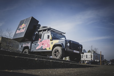 A rugged off-road vehicle with a large Red Bull can on its back, outfitted for promotional events. The vehicle has prominent branding with the Red Bull logo and is equipped with enhanced suspension and off-road tires. The backdrop features an overcast sky and a few trees and buildings in the distance.