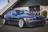 A modified blue BMW car is parked on a paved area in front of an industrial building. The car features large, stylish wheels and a lowered body kit. The backdrop shows a garage door with multiple red signs and neatly trimmed bushes around the area.