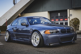 A modified blue BMW car is parked on a paved area in front of an industrial building. The car features large, stylish wheels and a lowered body kit. The backdrop shows a garage door with multiple red signs and neatly trimmed bushes around the area.