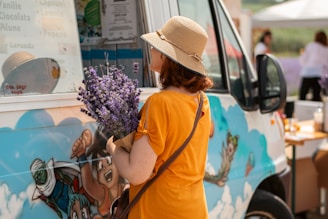 A woman wearing a straw hat and an orange shirt is holding a bouquet of lavender while standing next to a colorful food truck decorated with cartoon graphics. She appears to be reading the menu displayed on the truck's window.