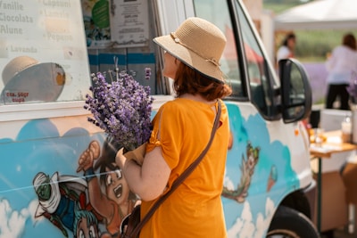 A woman wearing a straw hat and an orange shirt is holding a bouquet of lavender while standing next to a colorful food truck decorated with cartoon graphics. She appears to be reading the menu displayed on the truck's window.