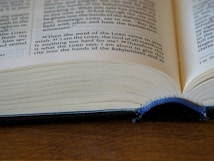 An open book with visible text on its pages, lying on a wooden surface. The page is slightly curved, with a blue bookmark cord hanging down.