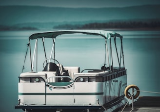 A pontoon boat is docked on calm, reflective water with lush, green hills visible in the background. The boat has white cushioned seats and a canopy for shade, with a serene and peaceful atmosphere.