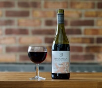 A bottle of red wine labeled McGuigan Family Release Shiraz is placed next to a filled wine glass on a wooden surface. The background consists of a rustic brick wall with a warm, earthy tone.