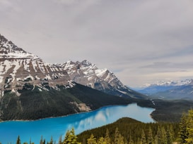 A stunning landscape featuring a turquoise glacial lake surrounded by dense evergreen forests. Snow-capped mountains rise majestically in the background under a cloudy sky.