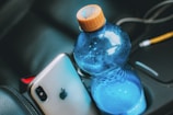 A smartphone with a silver back featuring a prominent logo lies next to a blue plastic water bottle with an orange cap, placed in a car's cup holder. The interior of the car is visible, with parts of the seat and console in the background.