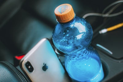 A smartphone with a silver back featuring a prominent logo lies next to a blue plastic water bottle with an orange cap, placed in a car's cup holder. The interior of the car is visible, with parts of the seat and console in the background.