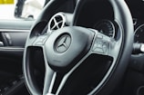 Close-up view of a car steering wheel featuring the brand's emblem at the center, with integrated control buttons on either side. The background includes dashboard elements like air vents and part of an instrument panel.