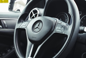 Close-up view of a car steering wheel featuring the brand's emblem at the center, with integrated control buttons on either side. The background includes dashboard elements like air vents and part of an instrument panel.