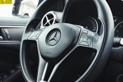 Close-up view of a car steering wheel featuring the brand's emblem at the center, with integrated control buttons on either side. The background includes dashboard elements like air vents and part of an instrument panel.