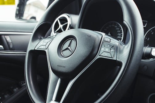 Close-up view of a car steering wheel featuring the brand's emblem at the center, with integrated control buttons on either side. The background includes dashboard elements like air vents and part of an instrument panel.