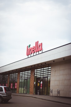 A storefront with a large sign displaying the name 'Linella' in red letters. The building features glass doors and windows, with red promotional signs displayed on them. There is a car parked in front of the entrance. The architecture includes a mix of white and beige panels, and the sky is overcast.