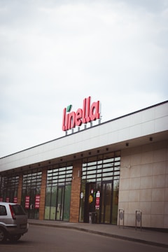 A storefront with a large sign displaying the name 'Linella' in red letters. The building features glass doors and windows, with red promotional signs displayed on them. There is a car parked in front of the entrance. The architecture includes a mix of white and beige panels, and the sky is overcast.