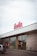 A storefront with a large sign displaying the name 'Linella' in red letters. The building features glass doors and windows, with red promotional signs displayed on them. There is a car parked in front of the entrance. The architecture includes a mix of white and beige panels, and the sky is overcast.