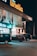 A nighttime street scene featuring a brightly lit storefront with signage that reads 'Linella'. Several parked cars are visible on the street, along with a large advertisement showing smartphones.