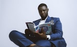 A person dressed in a blue suit, seated and engaged in reading a book titled 'Creative'. The suit is accessorized with a pocket square and a watch is visible on the wrist. The background is plain and light-colored, giving a minimalist appearance.