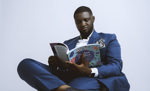 A person dressed in a blue suit, seated and engaged in reading a book titled 'Creative'. The suit is accessorized with a pocket square and a watch is visible on the wrist. The background is plain and light-colored, giving a minimalist appearance.