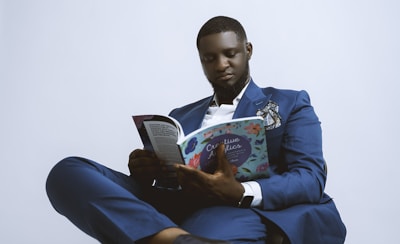 A person dressed in a blue suit, seated and engaged in reading a book titled 'Creative'. The suit is accessorized with a pocket square and a watch is visible on the wrist. The background is plain and light-colored, giving a minimalist appearance.