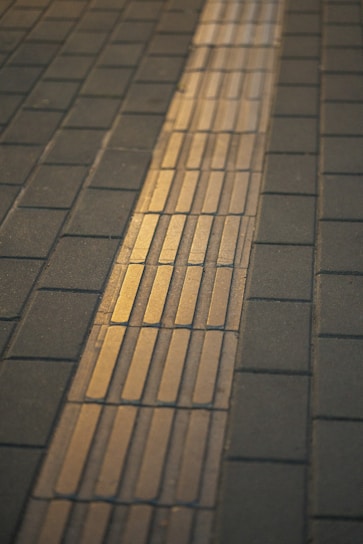 A sidewalk with a yellow tactile paving strip running through the middle, designed for visually impaired individuals. The paving consists of concrete tiles with a rectangular pattern, set against a darker background of regular paving stones.