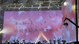 A stage performance with musicians seated under a large screen displaying an inspirational text in Chinese. Bright lighting and falling confetti add to the dynamic atmosphere. A stage crane is visible on the right side.