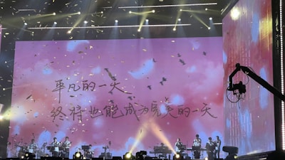 A stage performance with musicians seated under a large screen displaying an inspirational text in Chinese. Bright lighting and falling confetti add to the dynamic atmosphere. A stage crane is visible on the right side.