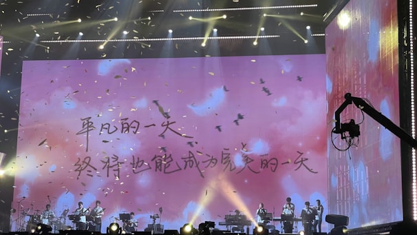 A stage performance with musicians seated under a large screen displaying an inspirational text in Chinese. Bright lighting and falling confetti add to the dynamic atmosphere. A stage crane is visible on the right side.