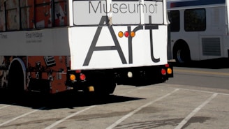 The back of a bus displays large black letters on a white background spelling out 'Museum of Art.' The bus features colorful artwork on its sides, and is parked in a parking lot under a clear sky. Another vehicle, likely another bus, is visible to the right.