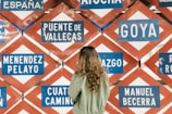 A person with long hair wearing a green jacket is standing in front of a wall filled with various blue and white signboards displaying names such as Puente de Vallecas, Goya, and Menendez Pelayo. The signs have a red and white diamond-shaped pattern as a background.
