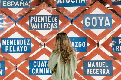 A person with long hair wearing a green jacket is standing in front of a wall filled with various blue and white signboards displaying names such as Puente de Vallecas, Goya, and Menendez Pelayo. The signs have a red and white diamond-shaped pattern as a background.