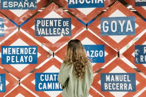 A person with long hair wearing a green jacket is standing in front of a wall filled with various blue and white signboards displaying names such as Puente de Vallecas, Goya, and Menendez Pelayo. The signs have a red and white diamond-shaped pattern as a background.