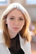 A young woman with light brown hair and freckles gazes directly at the camera. She is wearing a black top with a white outer layer, set against a softly focused background.