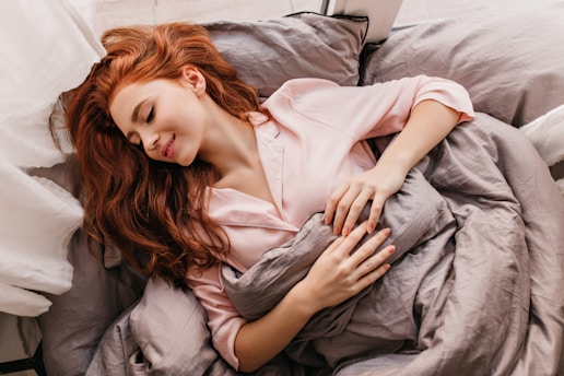 A person with long red hair, wearing a light pink pajama top, is peacefully sleeping on a bed with gray sheets. The scene is bright and calming, with soft natural light coming through what appears to be a window.