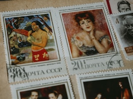 A collection of vintage postage stamps featuring artworks and portraits. One stamp displays a person in a vibrant red garment, surrounded by natural scenery. Another features a woman with red hair, adorned in an elegant dress, posing thoughtfully. Additional stamps feature monochromatic portraits of individuals from history.