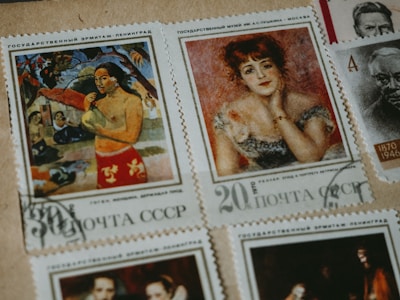 A collection of vintage postage stamps featuring artworks and portraits. One stamp displays a person in a vibrant red garment, surrounded by natural scenery. Another features a woman with red hair, adorned in an elegant dress, posing thoughtfully. Additional stamps feature monochromatic portraits of individuals from history.